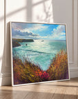 A framed coastal landscape painting leaning against a white paneled wall on a light wood floor, showing turquoise water, dramatic clouds, and grassy foreground in warm autumn tones.