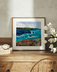 Framed Cornwall artwork of a coastal scene on a wooden dresser with flowers and a basket.