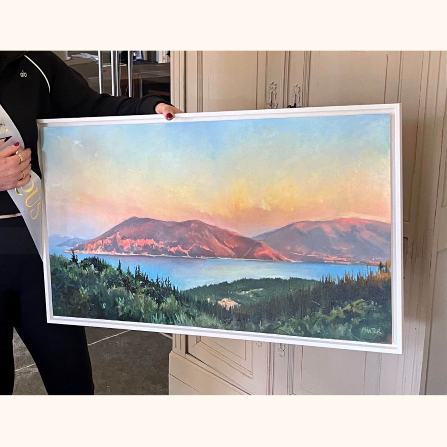 Mother holding a framed landscape commissioned painting as a birthday gift from her family of theview from their holiday retreat in front of Greek island mountains and and sunset lit sea
