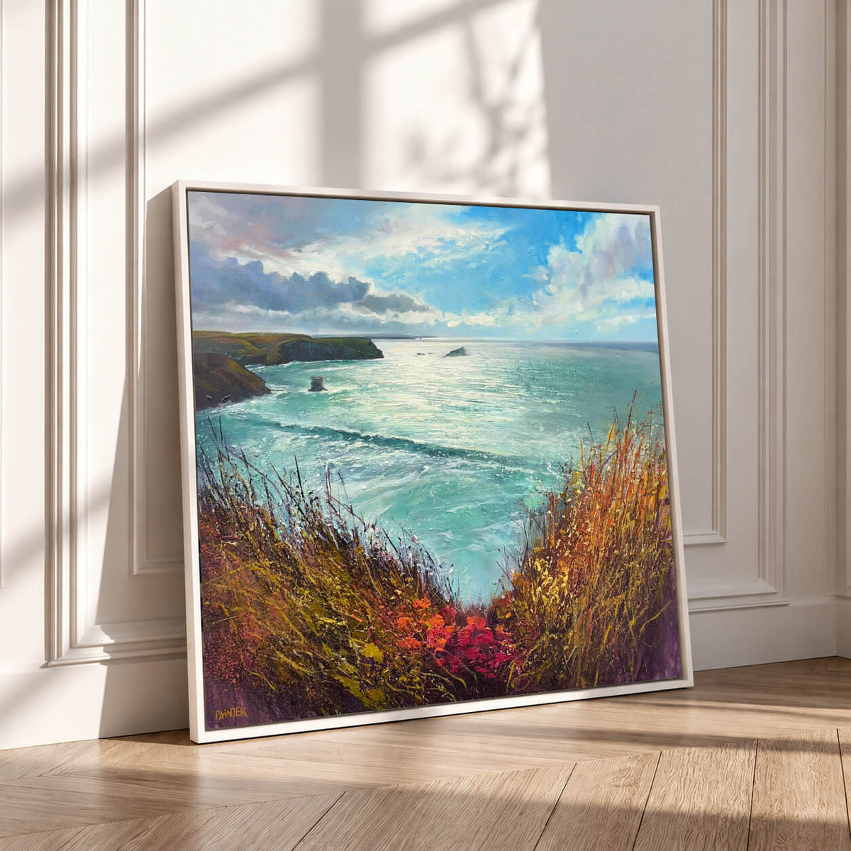 A framed coastal landscape painting leaning against a white paneled wall on a light wood floor, showing turquoise water, dramatic clouds, and grassy foreground in warm autumn tones.