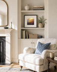 Atmospheric sunset landscape fine art print styled above a classic styled bookcase beside an armchair and fireplace, creating a cosy luxury reading nook interior. 