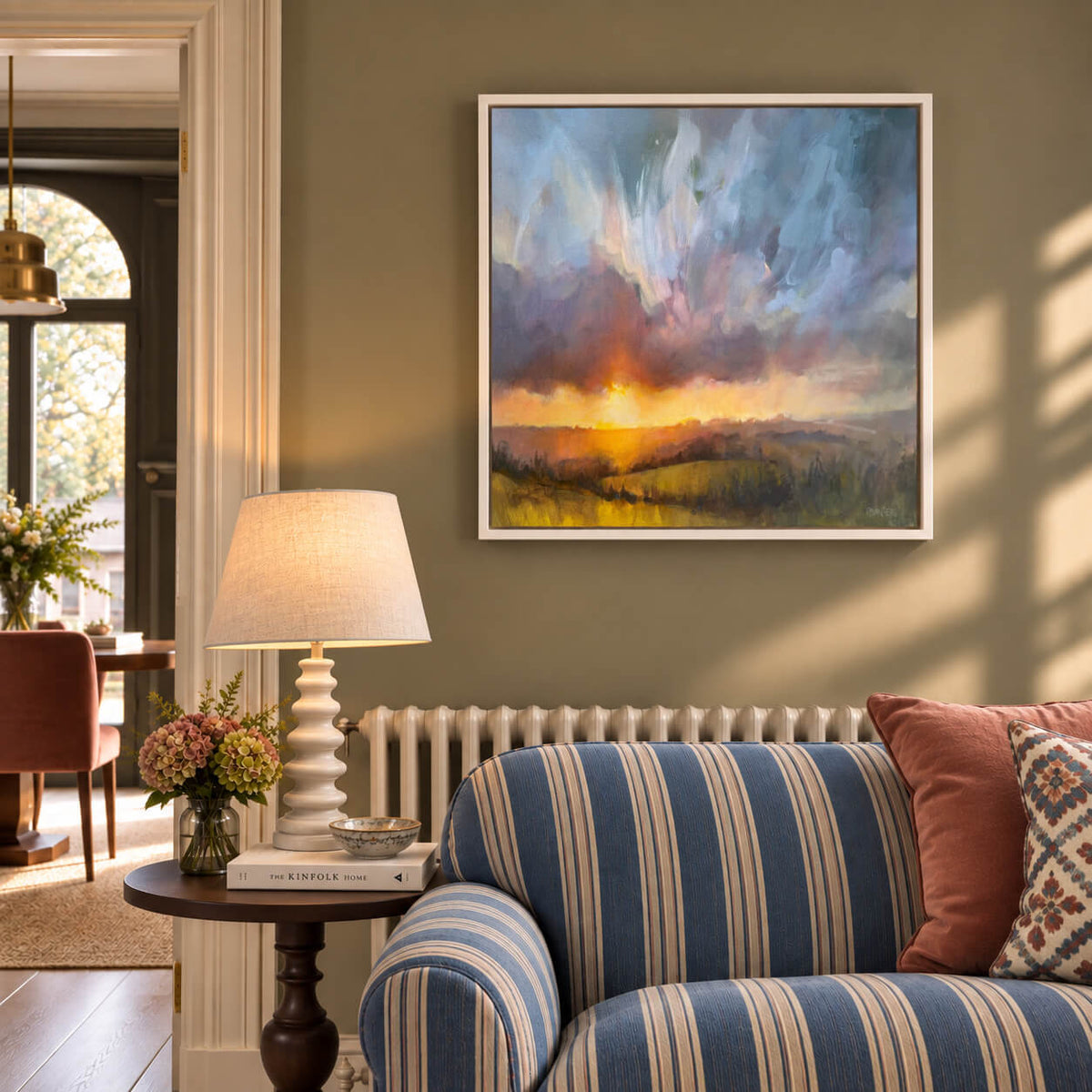 A framed landscape painting of a sunset over rolling hills, displayed on a sage green wall above a blue and cream striped sofa in a styled country interior living room.