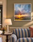 A framed landscape painting of a sunset over rolling hills, displayed on a sage green wall above a blue and cream striped sofa in a styled country interior living room.