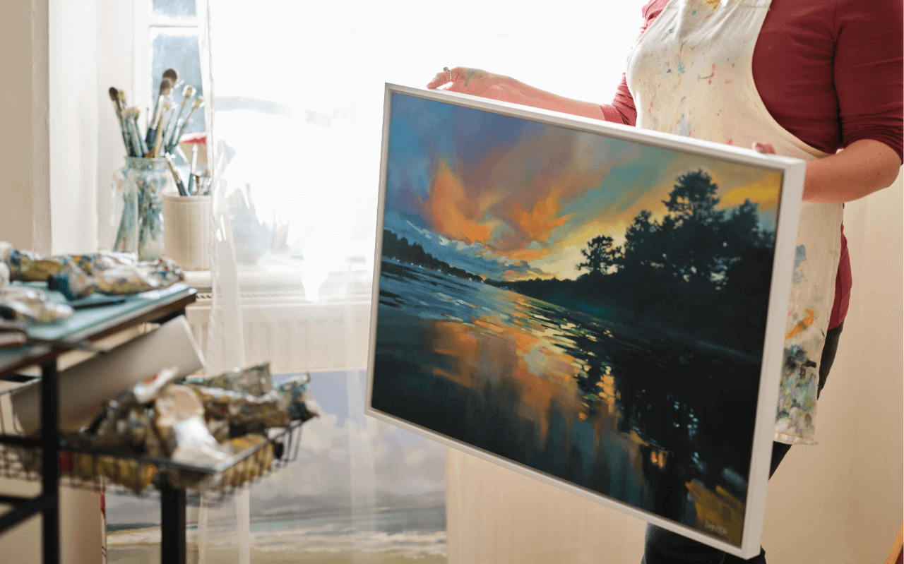 Rachel Painter holding a framed oil painting of a river landscape with trees and water in her artist studio in Cornwall