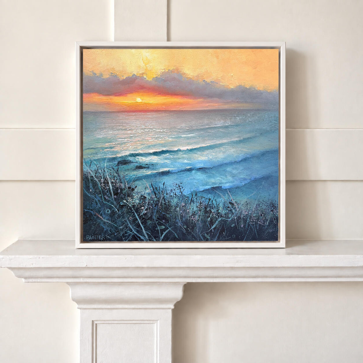 A framed square painting of a beach sunset with blue ocean waves, dark grasses in the foreground, and a warm orange and yellow sky, displayed on a white mantelpiece against a cream paneled wall.