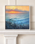 A framed square painting of a beach sunset with blue ocean waves, dark grasses in the foreground, and a warm orange and yellow sky, displayed on a white mantelpiece against a cream paneled wall.