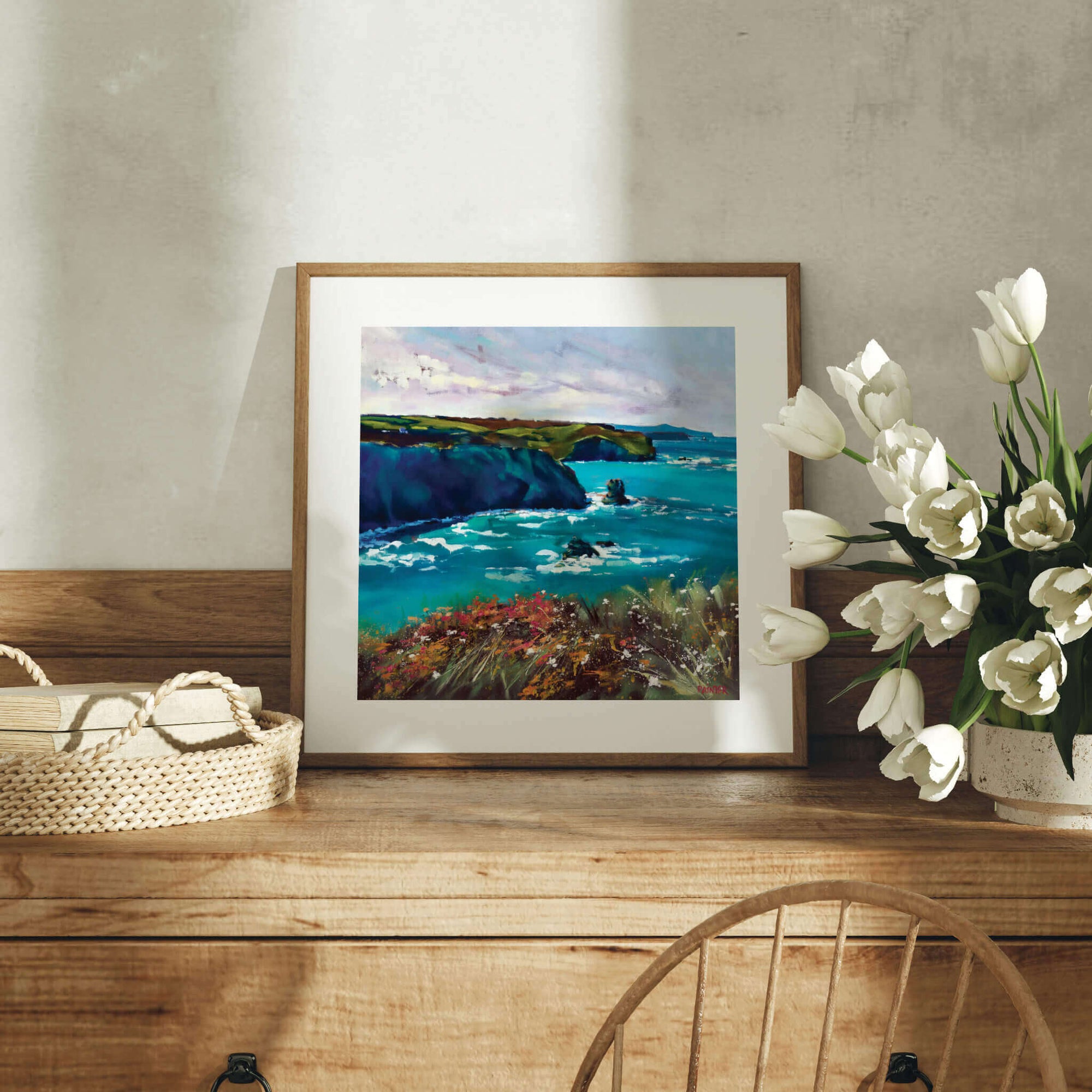 Framed Cornwall artwork of a coastal scene on a wooden dresser with flowers and a basket.